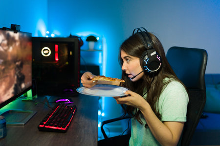 Hungry young woman with headphones eating a slice of pizza while playing a videogame on a gaming PC...
