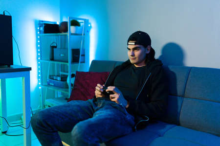 Hispanic man in his 20s lying on the couch in his bedrom decorated with led lights and having fun while playing a video game with a remote controllerの写真素材
