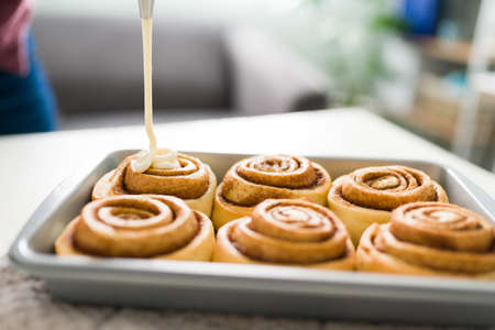 Woman using a piping nozzle to put the icing on the sweet rolls. Baked and glazed cinnamon rolls with white frosting on an oven trayの写真素材