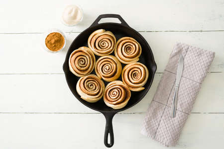 Flat lay of tasty cinnamon rolls fresh out of the oven on a white wooden table. Baked pastry fresh next to small bowls of sugar and cinnamon with a kitchen towel and a knifeの写真素材