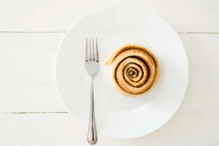 Flat lay of a delicious cinnamon roll on a white dessert plate with a fork. Top view of a sweet pastry ready to eat on the kitchen tableの写真素材