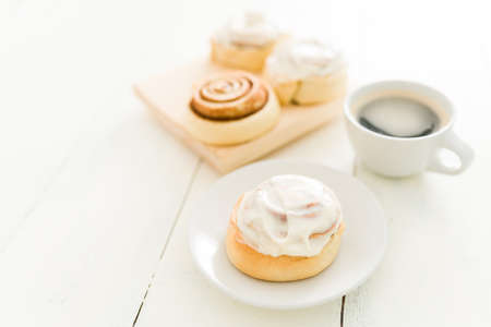 Enjoying a sweet snack in the afternoon. Beautiful glazed cinnamon roll with white frosting on a dessert plate with a hot cup of coffee on the kitchen tableの写真素材