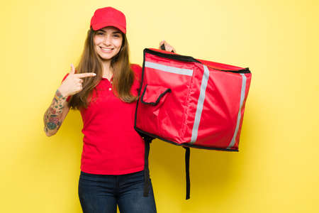 Latin woman making eye contact while pointing to her red backpack. Female courier ready to start delivering food to customers through a food delivery appの写真素材