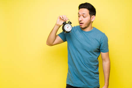 Stressed man in his 30s holding an alarm clock. Handsome man looking at the time and realizing the late hourの写真素材