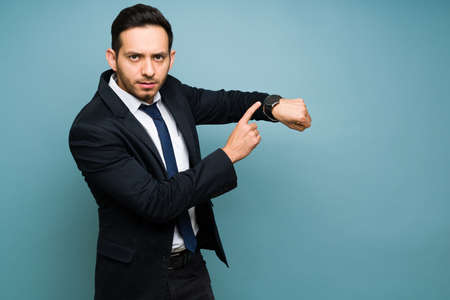 Angry attractive man wearing a formal suit and tie while pointing to the time in his watch. Hispanic...