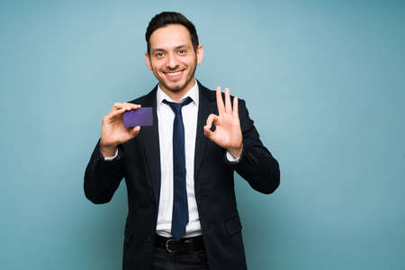 Happy man wearing a formal suit and a tie while holding a purple credit card. Attractive man doing the ok sign while showing his cardの写真素材