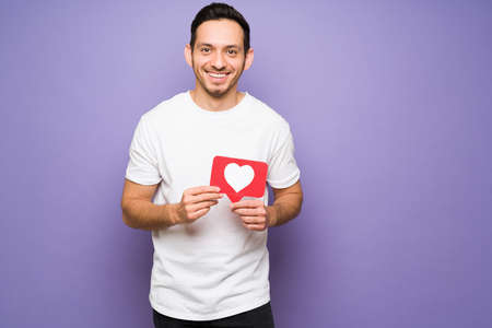Cheerful latin man with a beaming smile holding a like heart button. Attractive man using the like button on a social media appの写真素材