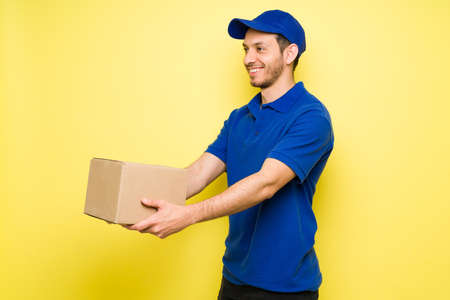 Happy latin man carrying a small package and making a delivery. Handsome male courier in a blue uniform smiling while workingの写真素材