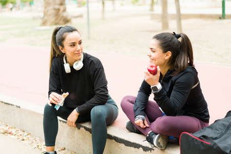 Happy and cheerful women talking about gossip while eating an apple and a power bar as a snack after doing a workout training in the parkの写真素材