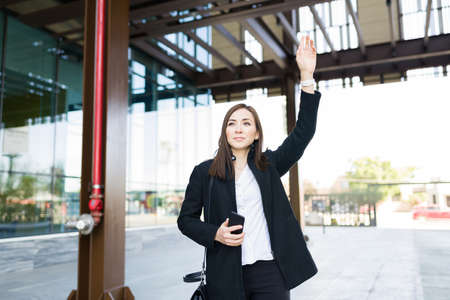 Attractive caucasian woman calling a taxi cab while holding her smartphone outside her modern office building in the cityの写真素材
