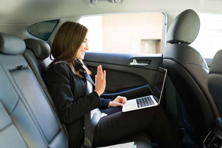 Successful business woman smiling and waving hello during a work video call on her laptop while sitting in the back of a car on her way to the office in the cityの写真素材