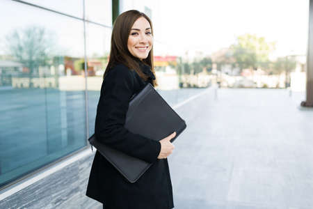 Female entrepreneur carrying a portfolio and walking outside a modern building. Smiling businesswoman working as a manager in a stylish office buildingの写真素材