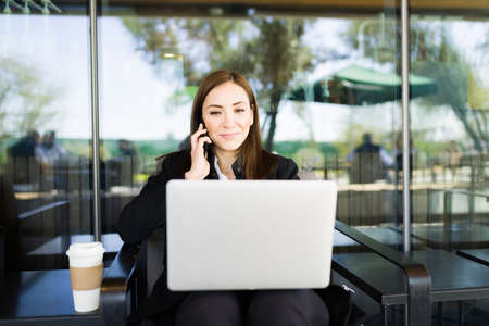 Busy business woman talking on the phone during a work call while typing on her laptop. Successful businesswoman drinking coffee and working on a coffee shopの写真素材