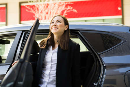 Successful entrepreneur getting out of a car and smiling while looking up to a building in the business district in the city. Happy businesswoman arriving at the office on a taxi cabの写真素材