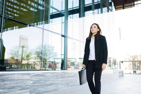 Attractive business woman walking to her city office carrying a portfolio. Professional businesswoman standing outside a modern building with a serious expressionの写真素材