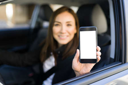 Happy woman holding her smartphone and smiling while working as a driver for a ride sharing mobile app. Caucasian woman showing the blank screen of her phoneの写真素材