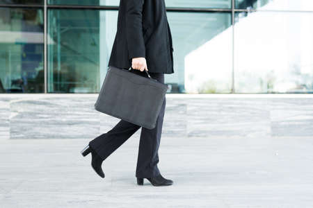 Close up of a caucasian business woman walking to her office outside a modern building in the city while carrying a black portfolioの写真素材