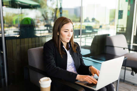 Busy business woman sitting on the patio of a coffee shop and typing on her laptop. Female entrepreneur working on the computer at a cafe while drinking coffeeの写真素材