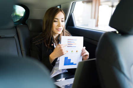 Smart businesswoman explaining a work report or analysis to her colleagues and coworkers during a online video call on her laptop while sitting in the back of a taxi cabの写真素材