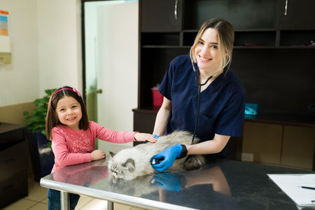 Beautiful professional veterinarian smiling and making eye contact while doing a medical exam on a white persian cat. Little girl owner calming down her pet at the vetの写真素材