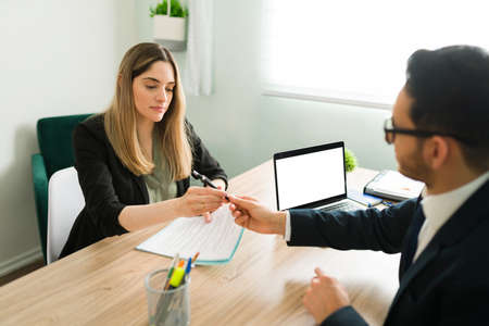Male lawyer giving a business contract and a pen to a caucasian woman sitting at his desk office. Female colleague about to sign work papers with her bossの写真素材