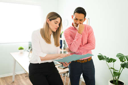 Beautiful woman pointing and holding work papers. Female coworker explaining a contract to a male hispanic colleague in the officeの写真素材