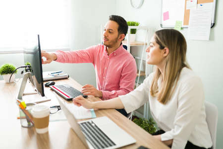 Handsome man speaking and showing a business presentation to a caucasian woman coworker while looking to the screen on the computer. Professional colleagues sitting at their deskの写真素材