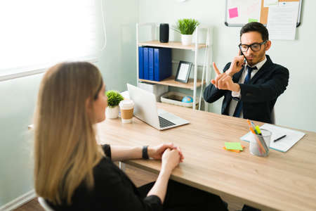 Handsome hispanic man wearing glasses and a suit talking on the phone with a client while telling a female caucasian coworker to hold on a minuteの写真素材