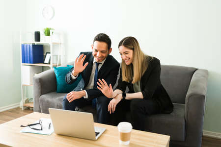 Smiling business partners waving and saying hello to coworkers and colleagues during a video call on a laptop. Business woman and man during an online meeting at the officeの写真素材