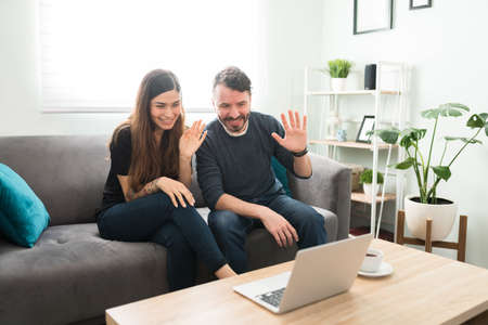 Attractive married couple at home smiling and waving hello to their happy friends and family during...