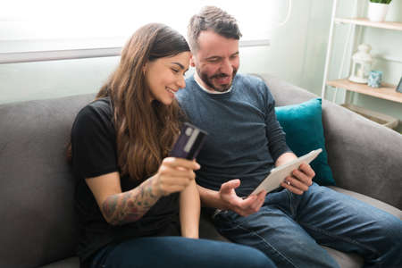 Beautiful young woman holding a credit card and a tablet to pay for the online shopping with her husband while sitting on the couchの写真素材
