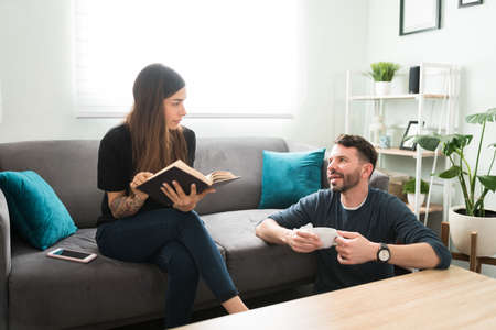 Handsome man talking about his day with his girlfriend while drinking a cup of coffee at home. Young wife holding a book and listening to her husbandの写真素材