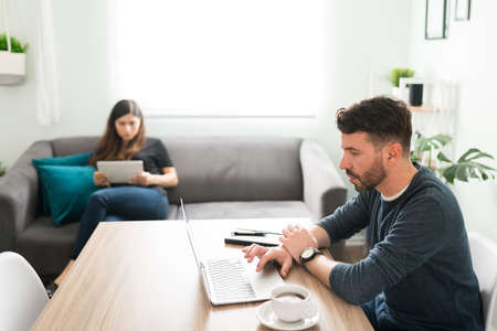 Handsome man sitting at the living room table and working on his laptop with his beautiful wife. Young woman using his tablet on the sofaの写真素材