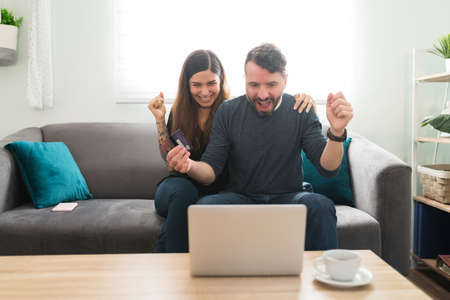 Excited hispanic couple paying with a credit card for their online shopping on a laptop. Surprised husband and wife feeling happy because of a big sale and discount onlineの写真素材
