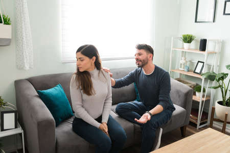 Attractive young woman ignoring her upset husband while sitting together on the sofa. Handsome boyfriend asking for forgivenessの写真素材