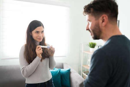 Happy young woman holding a pregnancy test and surprising her husband with positive results. Beautiful wife telling a latin man that she is pregnantの写真素材