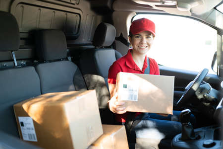 Excited young woman working as a delivery courier and sitting behind the wheel of a truck. Caucasian female worker choosing a package to deliver at a shipping addressの写真素材