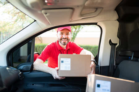 Excited male courier and delivery man holding a small package in the delivery van while standing...