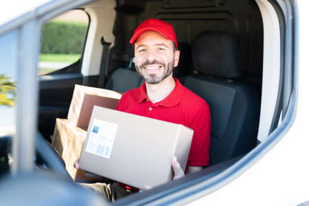 Happy male courier arriving to the shipping address in a delivery truck and holding a small package....