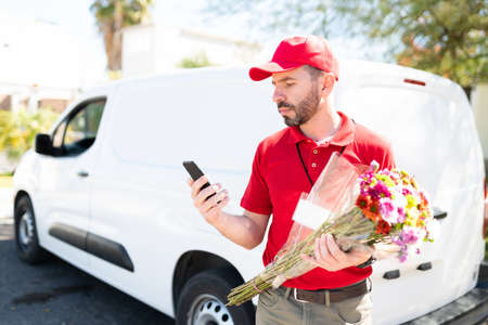 Hispanic delivery man in a red uniform reading the contact information of a customer in his smartphone to make a home delivery service. Male worker delivering a bouquet of flowersの写真素材