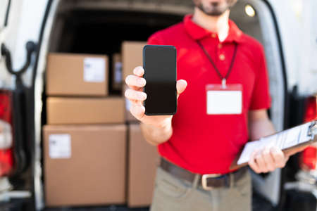 Delivery male courier showing the screen of a smartphone while delivering packages. Delivery man using a phone to read the customer information and shipping addressの写真素材