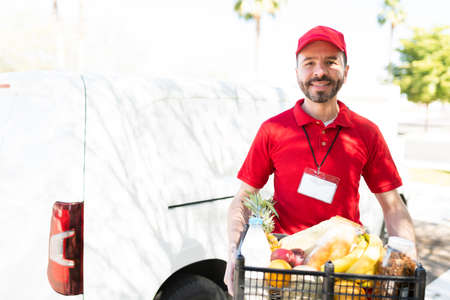 Portrait of a handsome hispanic man holding a box of fresh products and produce from the supermarket and ready to make a home delivery serviceの写真素材
