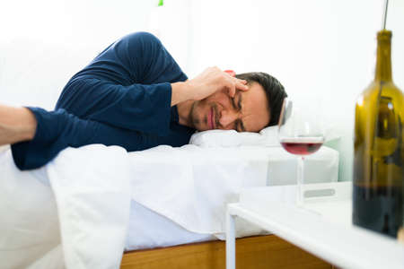 Handsome man waking up in his bedroom after drinking a lot of wine. Young man rubbing his head...