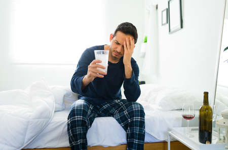 Sick man in pajamas sitting in bed after waking and about to drink a glass of water with an...