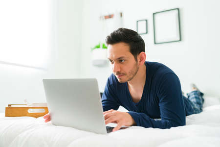 Attractive young man in pajamas lying on his stomach in bed while typing on a laptop next to a tray...