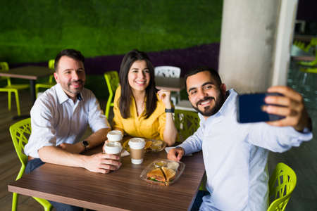 Attractive friends and co-workers taking a selfie with a smartphone while sitting together at a table in the food court and eating lunch after workの写真素材