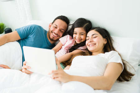 Happy young mom and dad laughing with her little daughter in the bedroom while watching comedy...