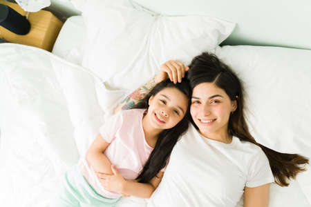 Portrait of a cute little girl and beautiful young mom cuddling in bed together after waking up in a...