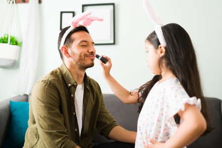 Playful little girl with bunny ears putting lipstick on her father's lips while playing dressing up at homeの写真素材