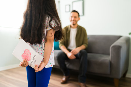 Celebrating a Happy Father's Day. Cute little girl hiding a handmade card behind her back and smiling to her dad sitting on the sofa at homeの写真素材